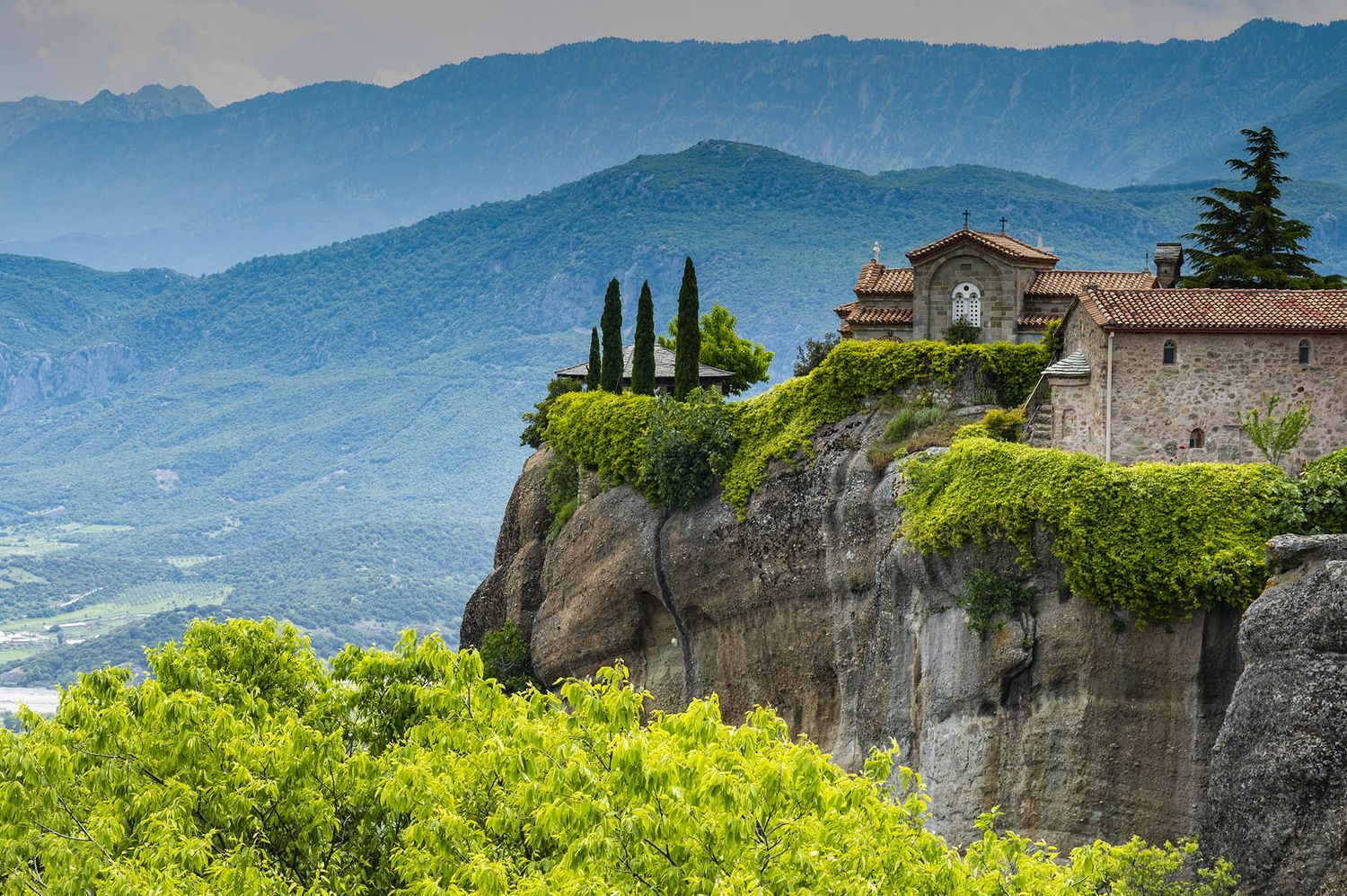 Panoramic view of historic Meteora monasteries perched on towering rock formations in Greece with misty mountains in background.