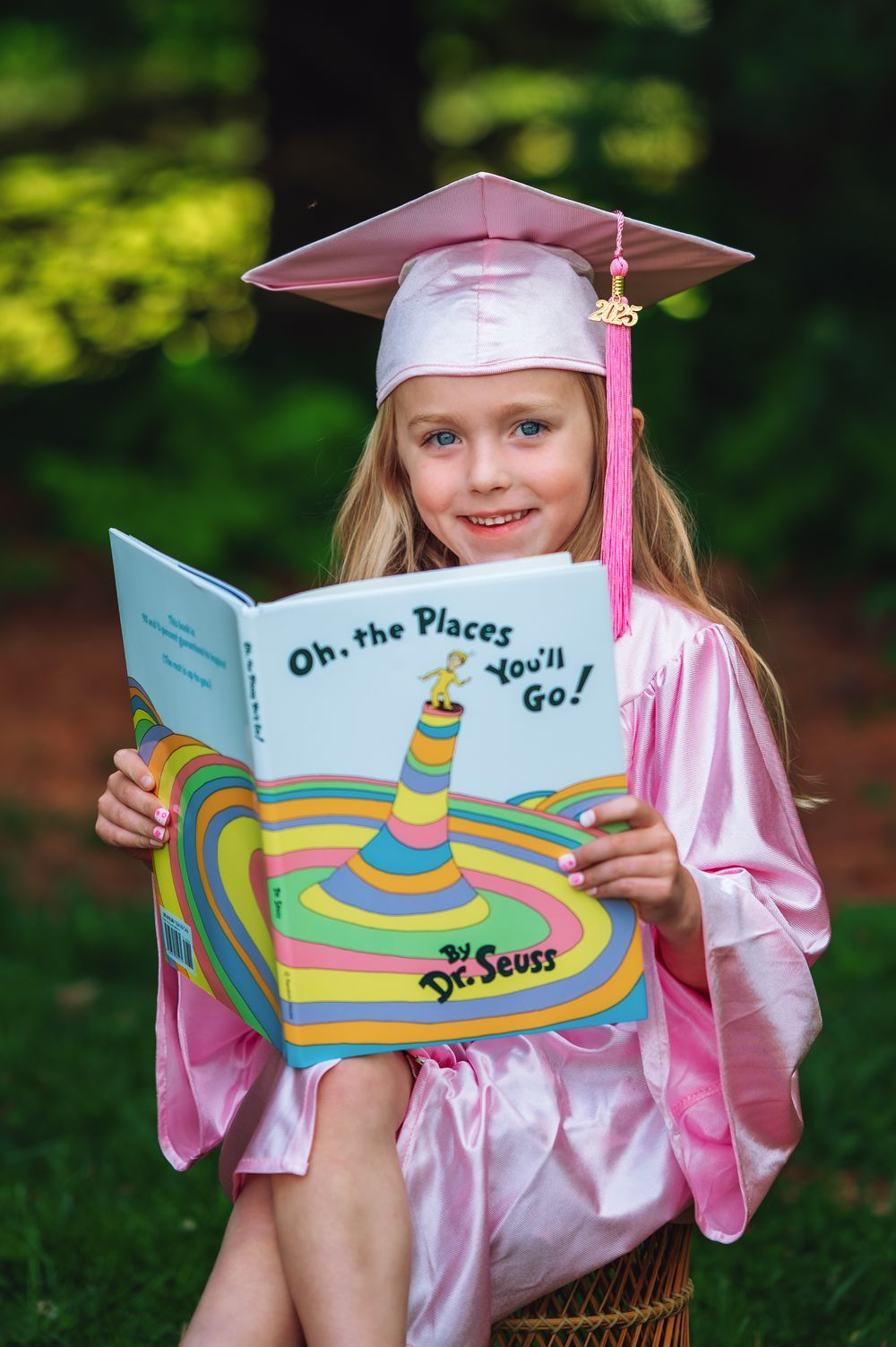 Graduate in pink cap and gown reading Dr. Seuss's 'Oh The Places You'll Go' book in outdoor setting.