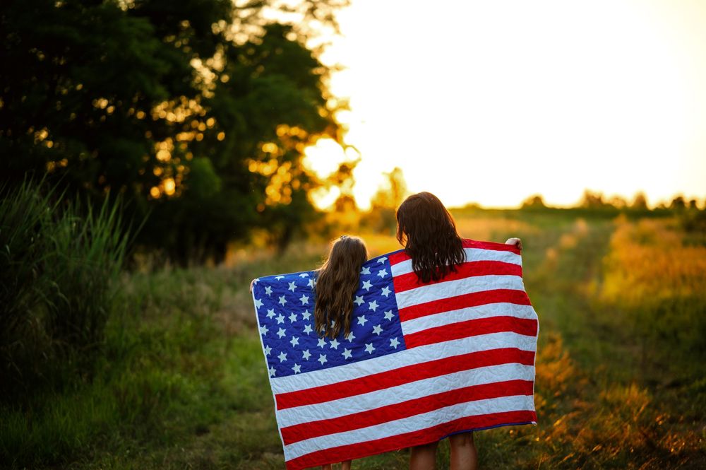 American flag waves against golden sunset while silhouetted trees frame scenic rural landscape at dusk.