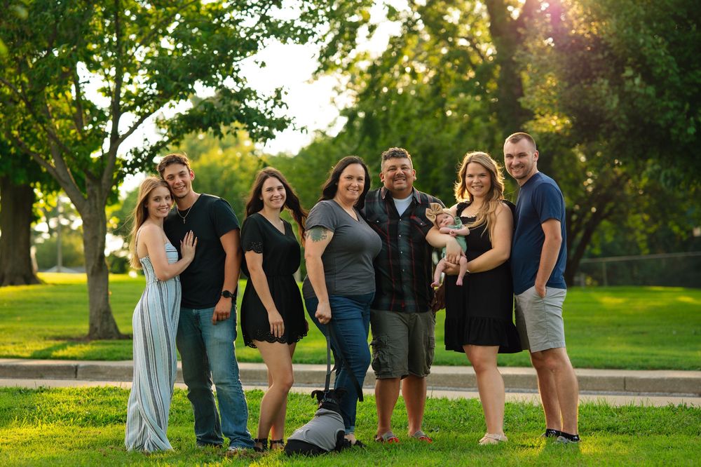People gather for outdoor family photos in a park during golden hour with trees and sunlight in the background.