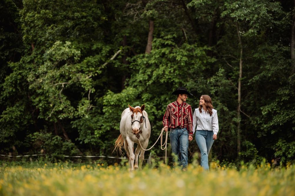 Couple walking a white horse through a lush green meadow surrounded by dense forest trees.