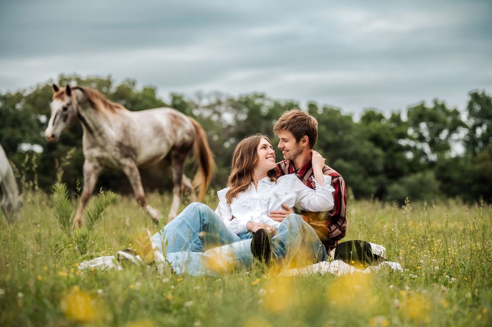 Couple sitting in a wildflower meadow with horses grazing behind them on a cloudy day.