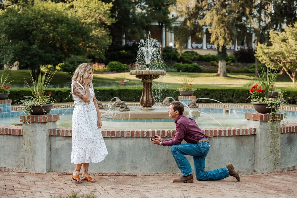 Man proposing on one knee to woman by ornate garden fountain surrounded by lush greenery and flowers.