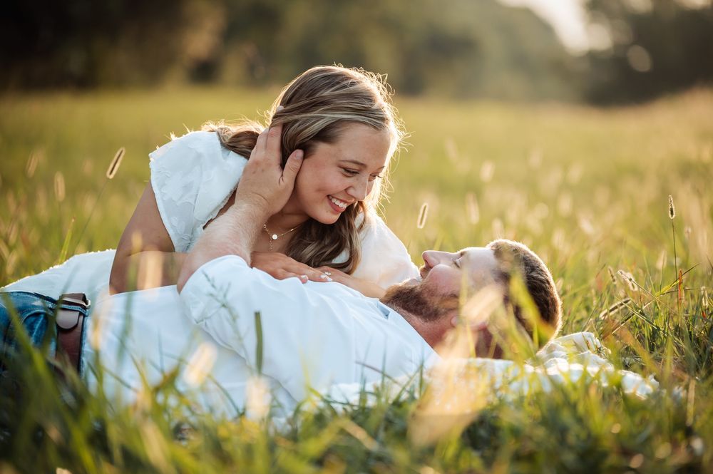 Couple smiling and laughing together while lying in a sunny green meadow, sharing a romantic outdoor moment.