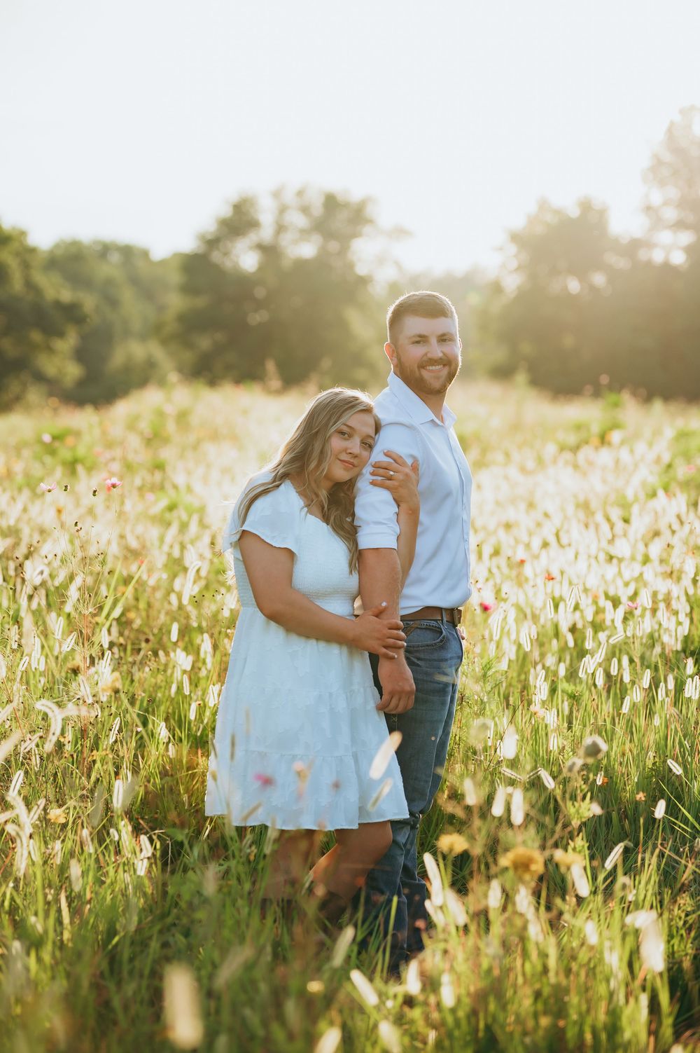 Couple embracing in a sunlit wildflower field, woman in light blue dress, man in white shirt and jeans, golden hour.