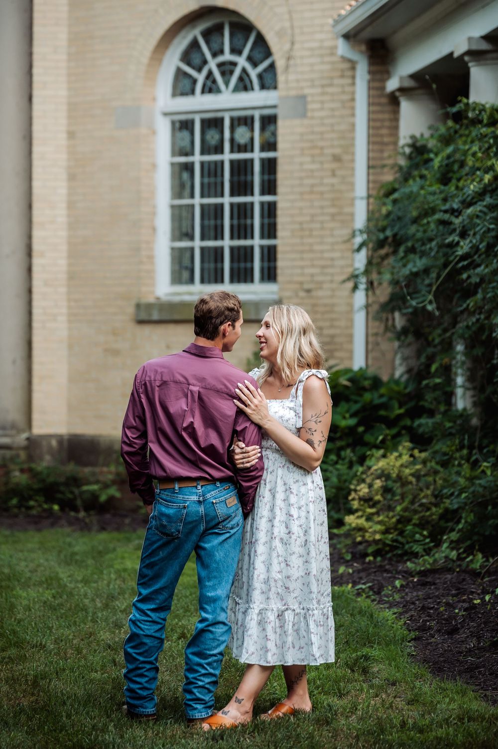 Couple in love standing on green lawn outside brick building, man in maroon shirt and jeans, woman in floral dress.