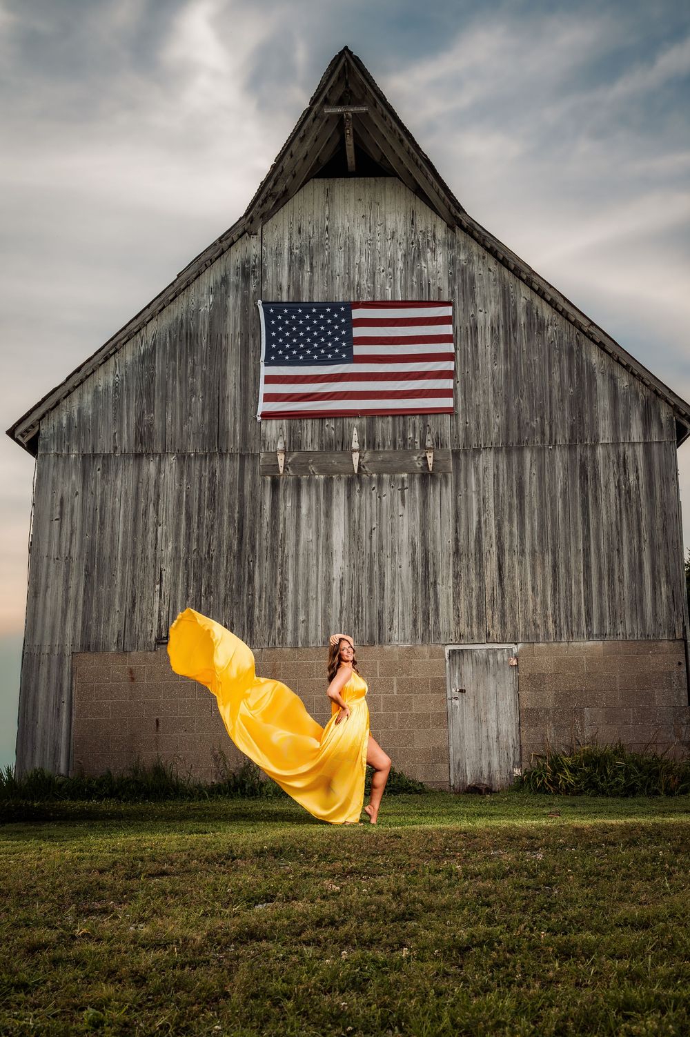Woman in flowing yellow dress poses before rustic barn with American flag on cloudy day.