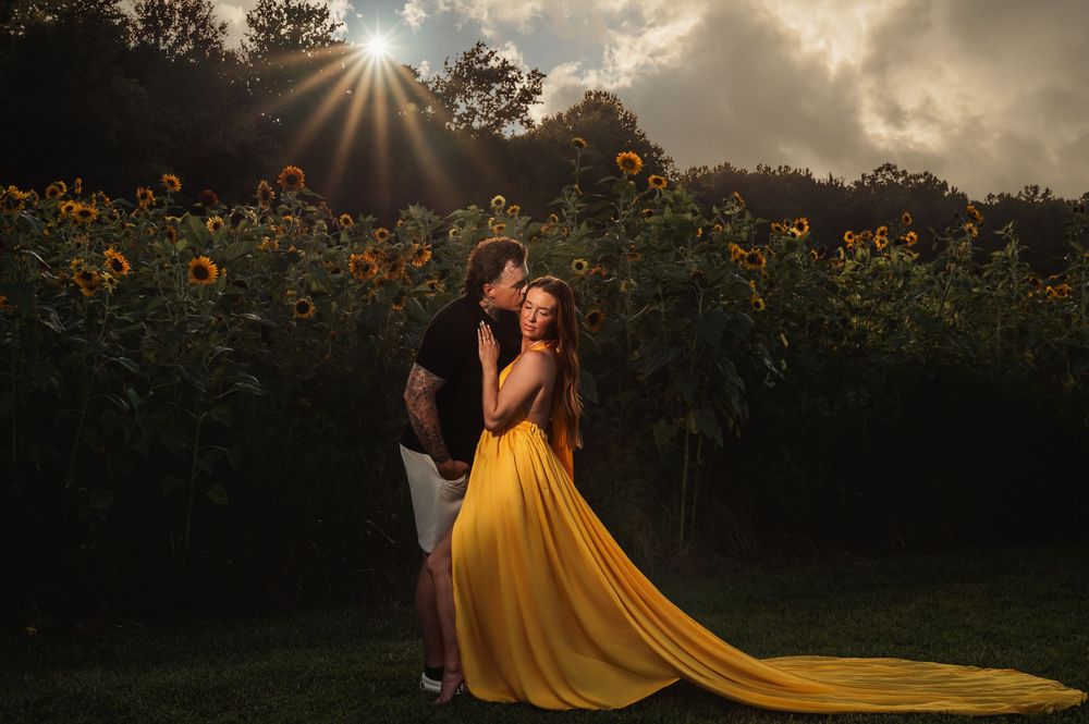 Couple embracing in a sunflower field at golden hour, woman in flowing yellow gown with dramatic sunburst sky.