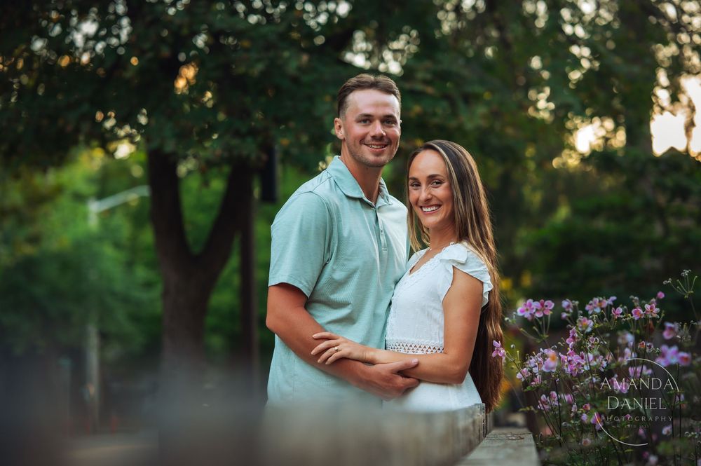 Couple embraces during outdoor photo session with blooming purple flowers and trees at sunset.