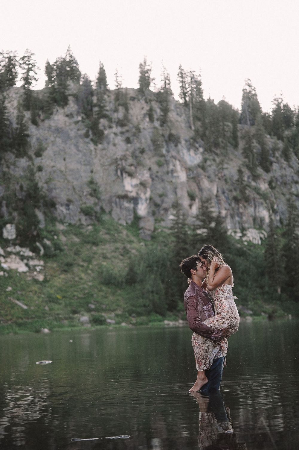 Couple embracing in shallow water with dramatic cliff face and forest in background.