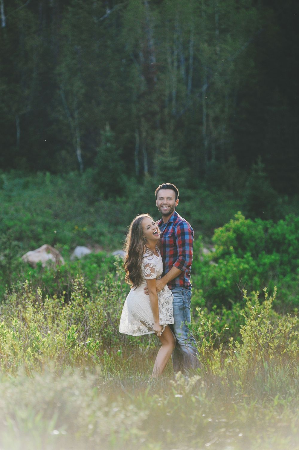 A couple shares playful moments in a meadow with lush forest in the background.