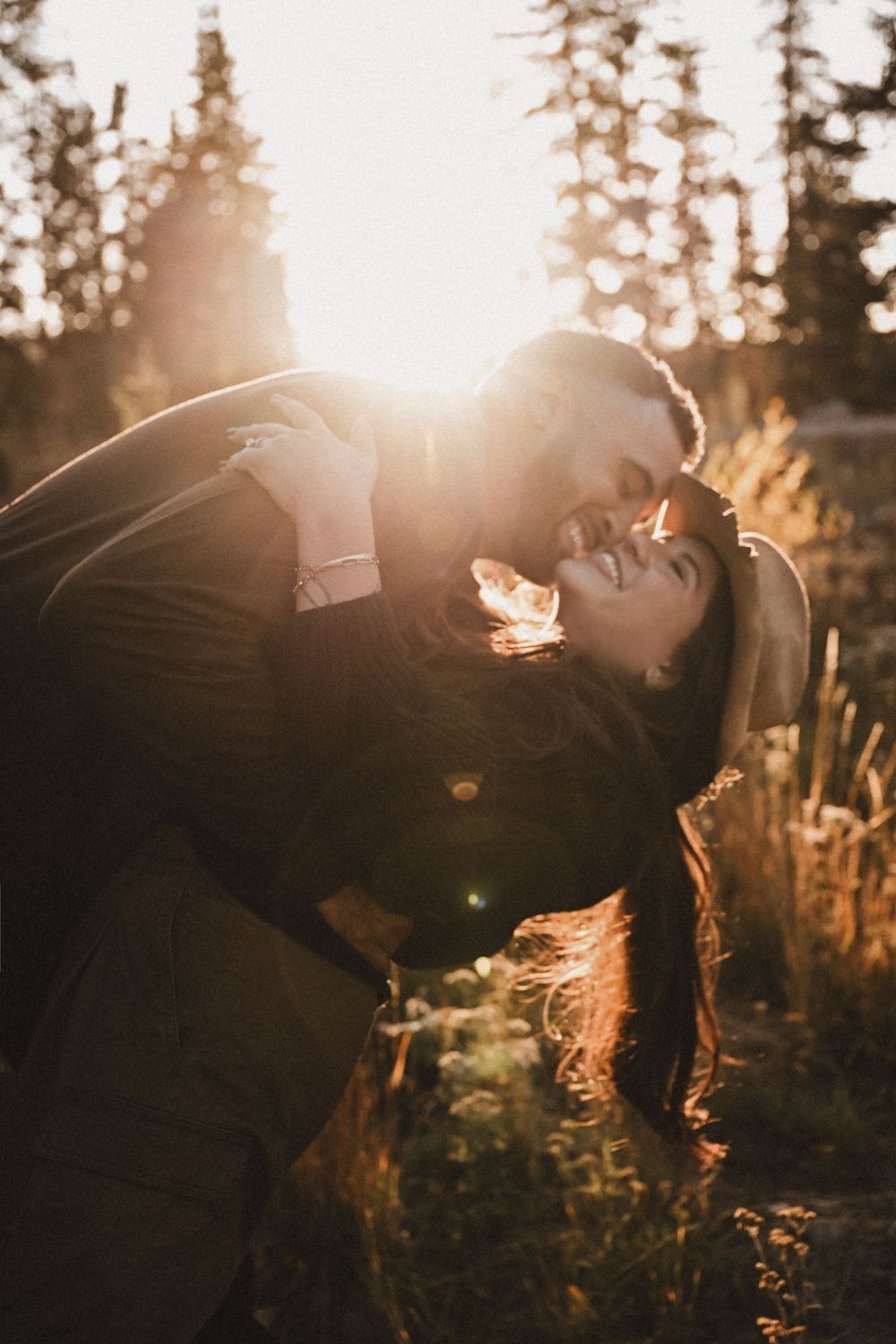 Romantic couple kissing in golden sunset light surrounded by evergreen trees and tall grass.