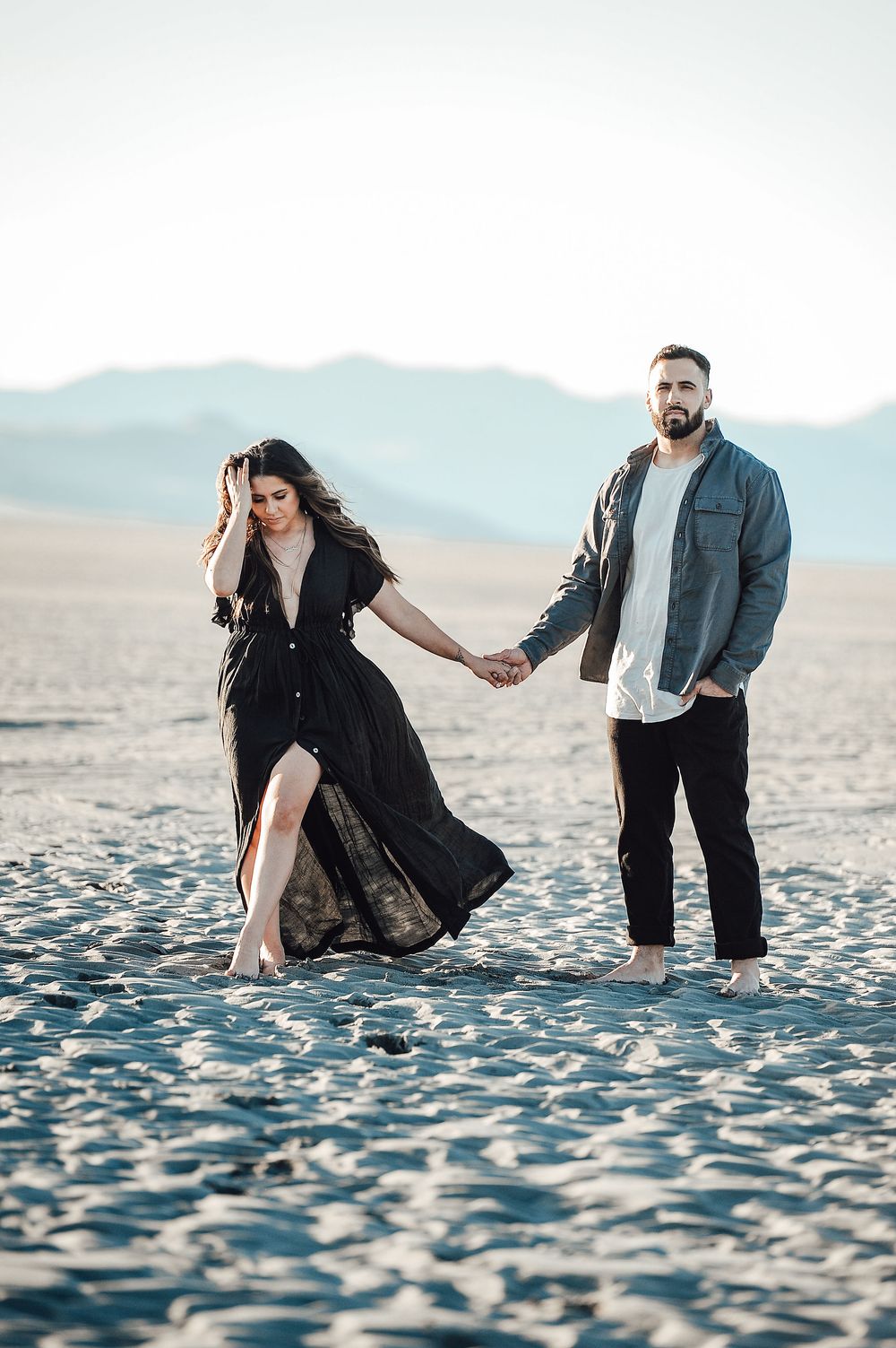 Playful couple dancing and moving together on sandy beach during outdoor photoshoot.