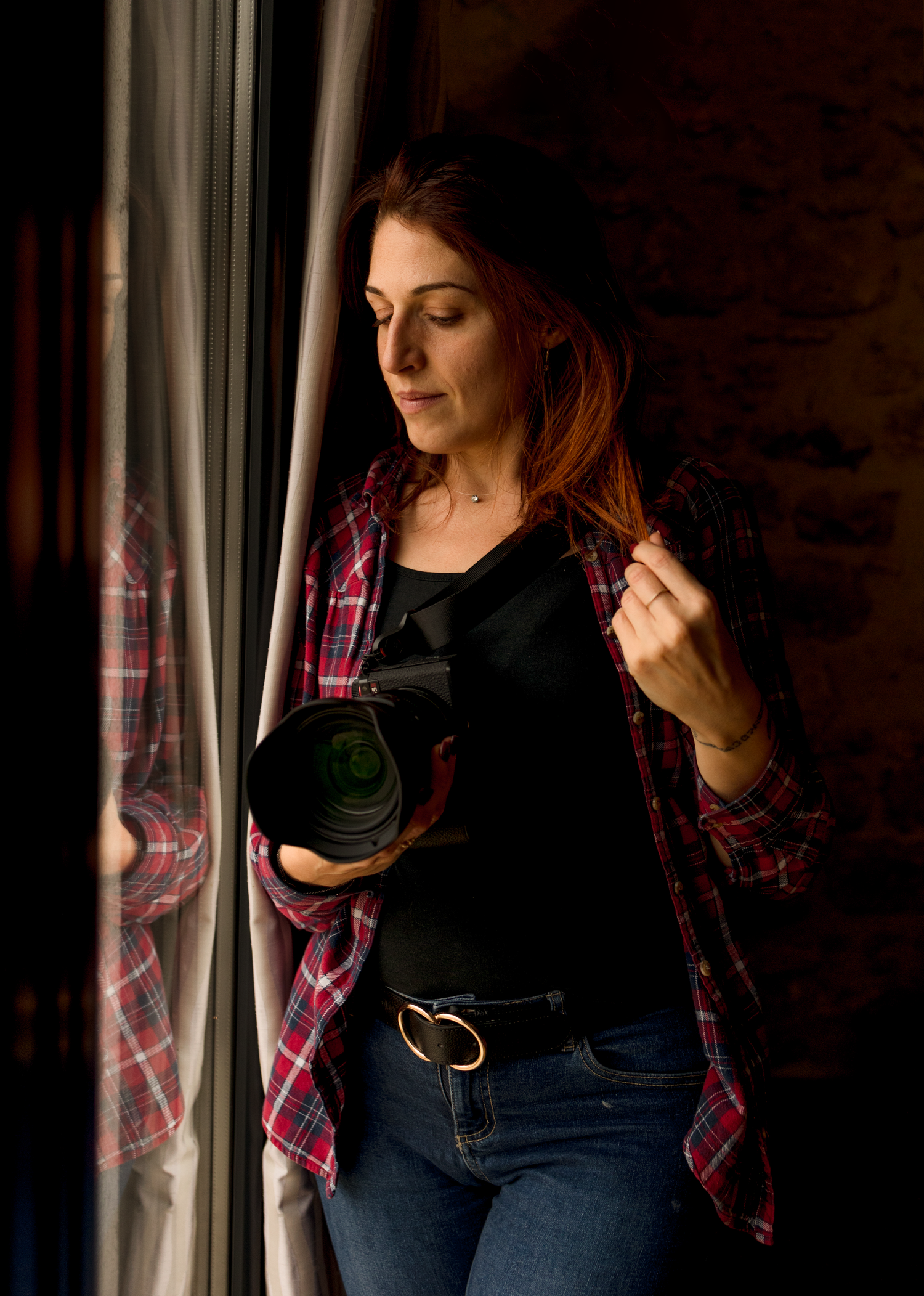 Thoughtful figure stands by window in plaid flannel shirt and jeans, holding camera in warm natural lighting.