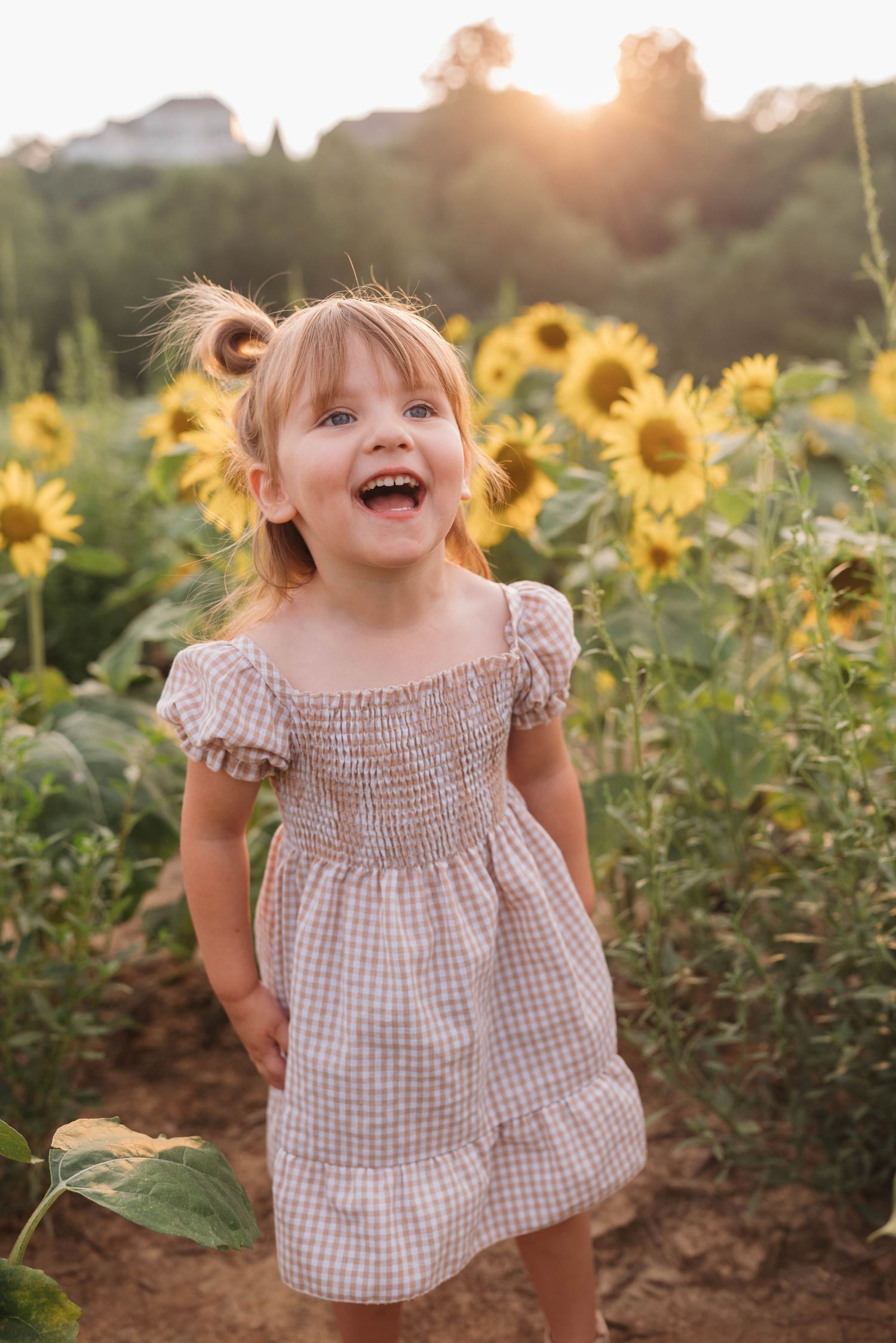 Child in gingham dress laughing joyfully in sunflower field at sunset.
