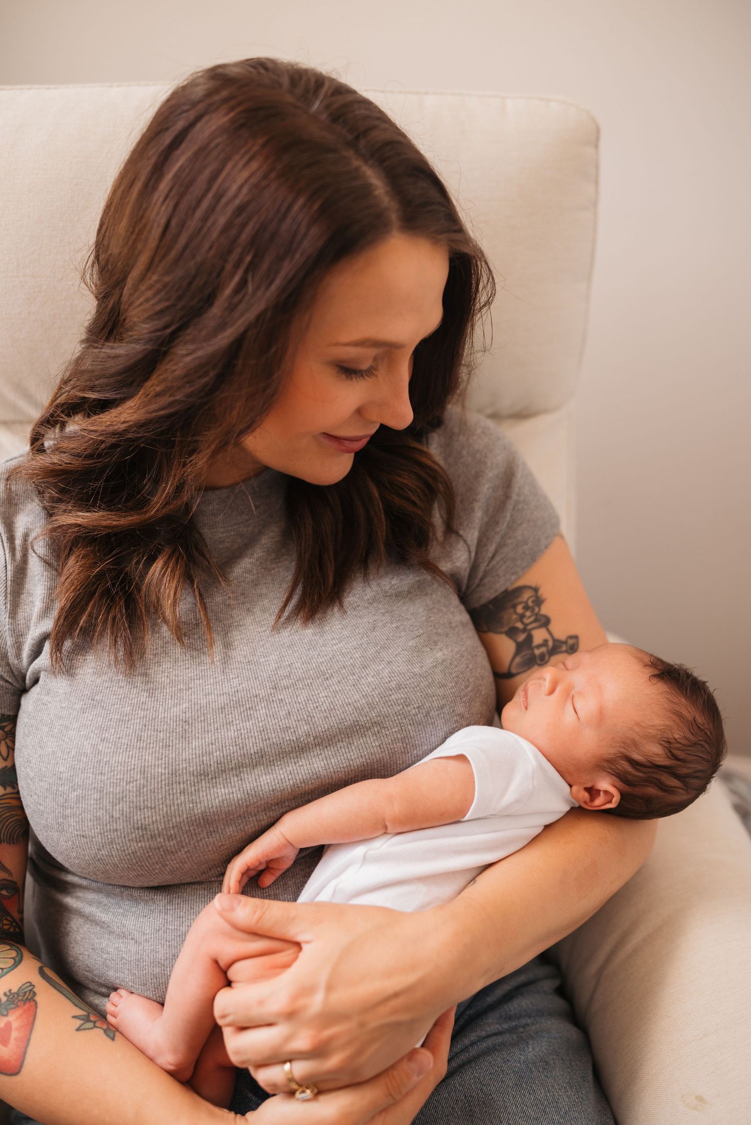 Mother in grey shirt tenderly holding sleeping newborn on white couch.