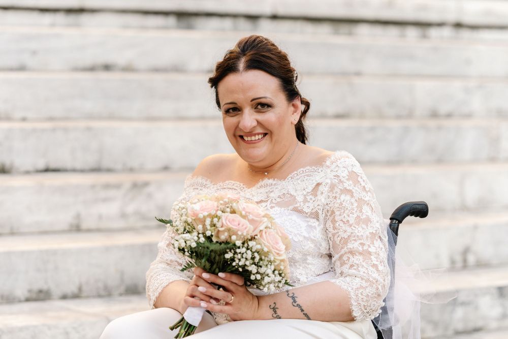A bride in a white lace dress with three-quarter sleeves sits on stone steps holding a delicate bouquet.