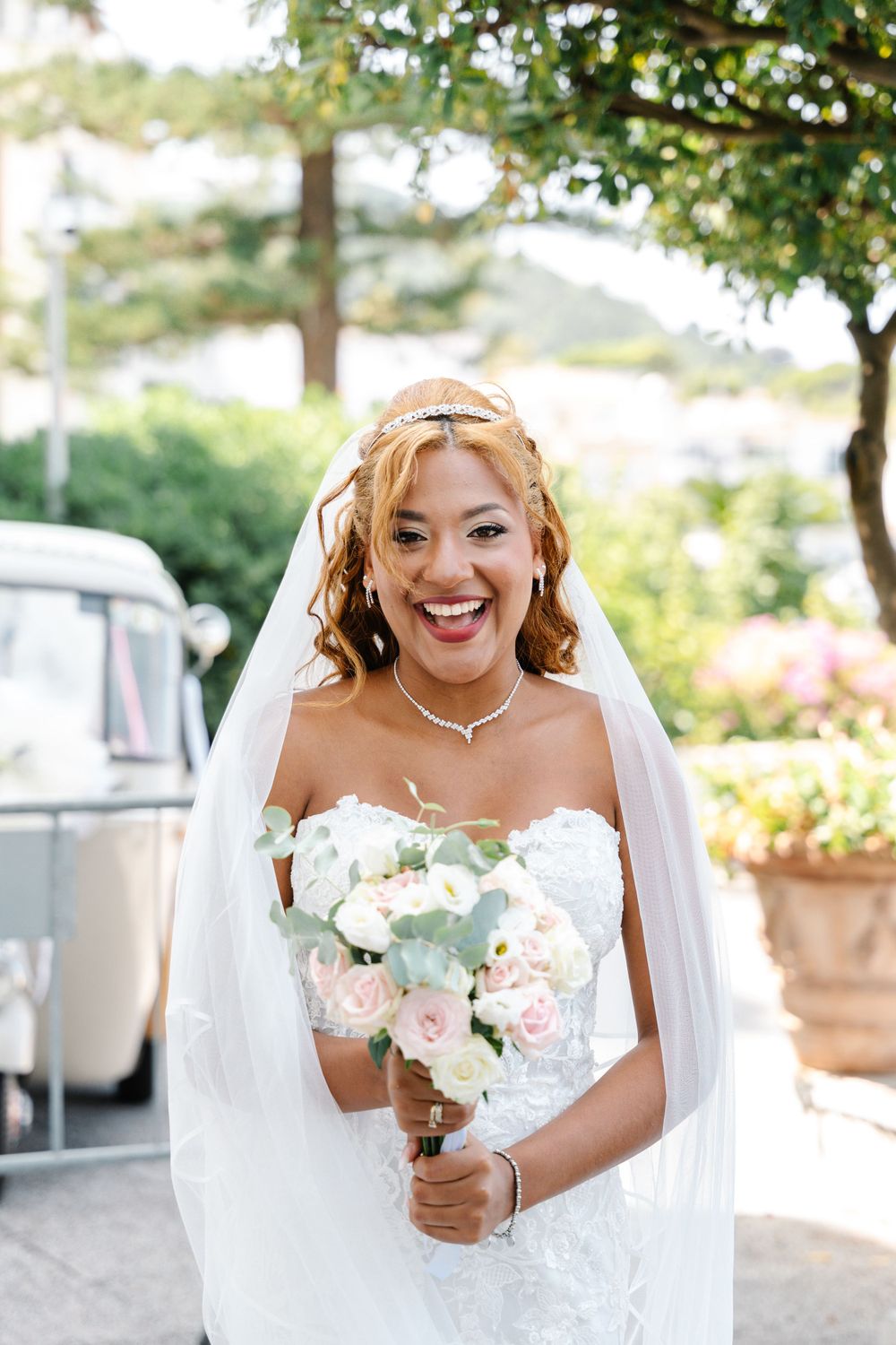 A bride in a strapless wedding dress holds a white and pink bouquet against a garden background.
