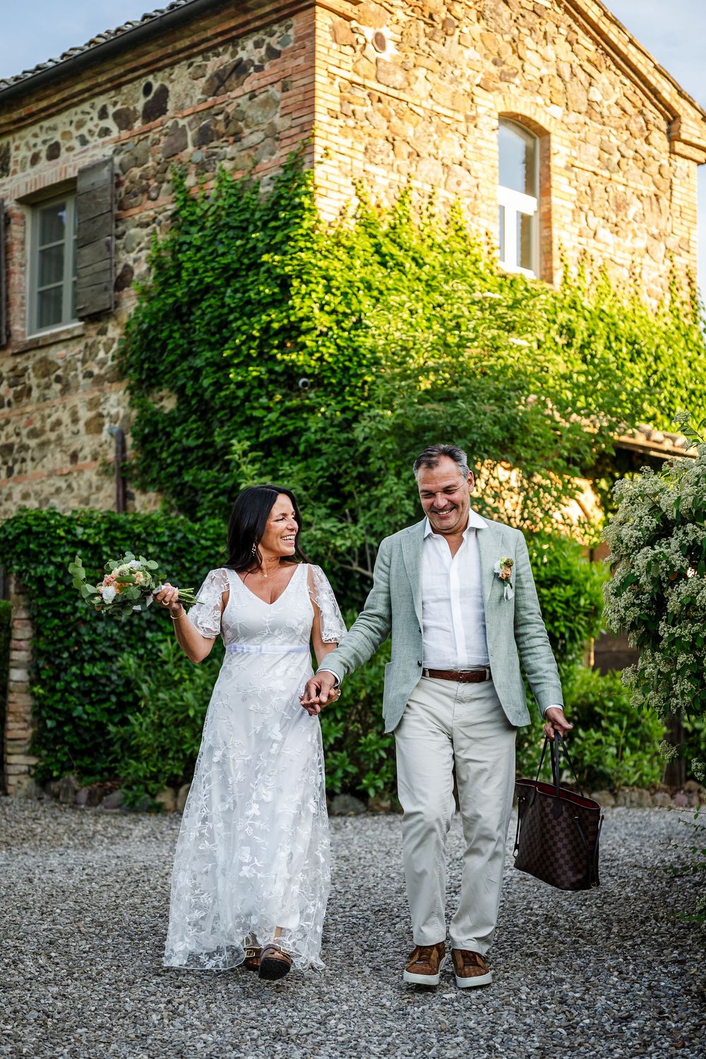A couple in wedding attire walking together outside a rustic stone building with ivy-covered walls in warm sunlight.