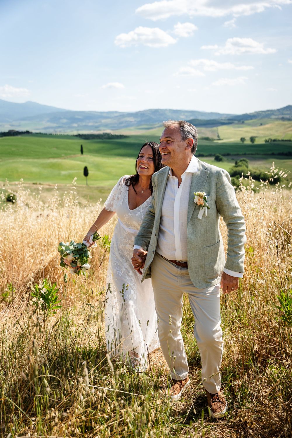 Newlyweds share laughter while walking through golden wheat fields with mountains in the distance.