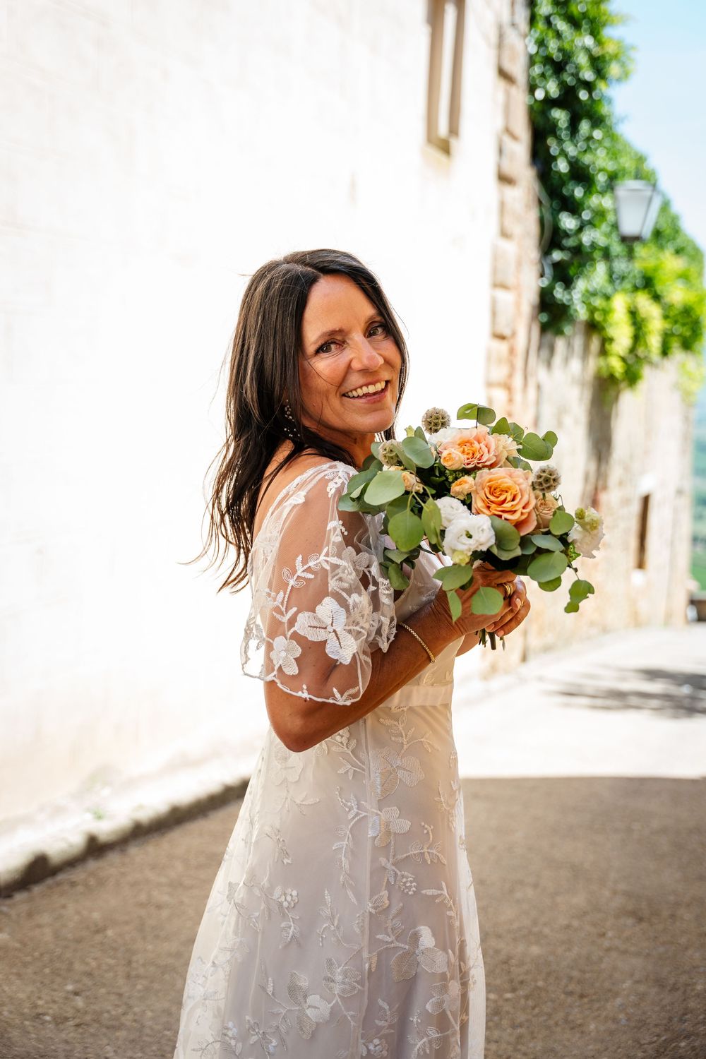 Bride in flowing lace dress twirls joyfully while holding a peach and white floral bouquet.