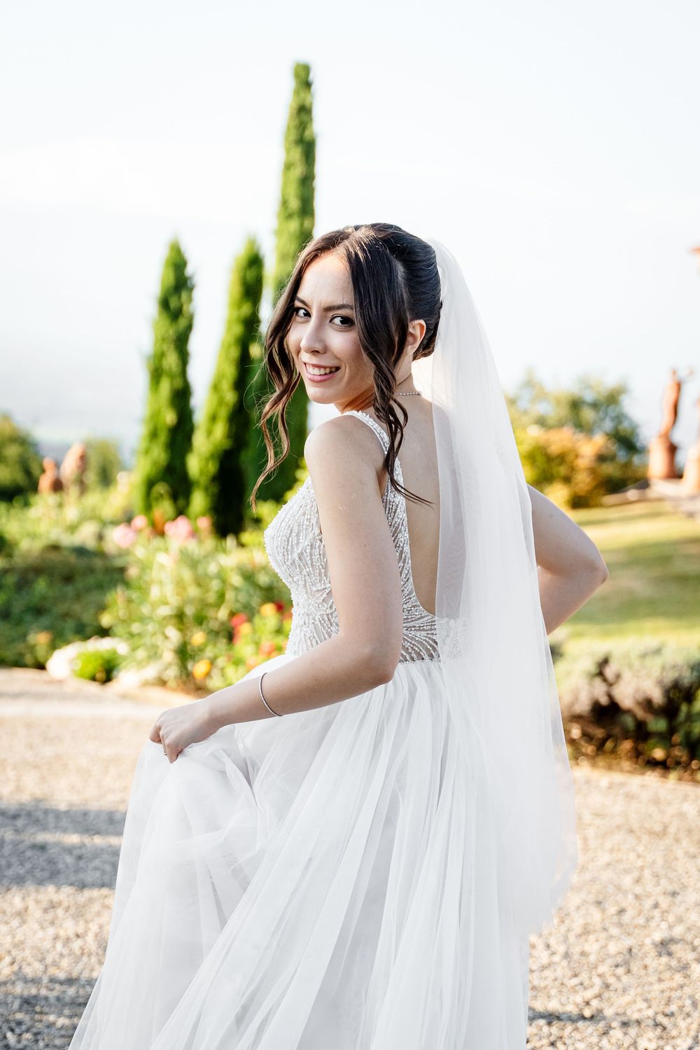 A bride in a flowing white gown twirls gracefully on a gravel path surrounded by lush greenery.