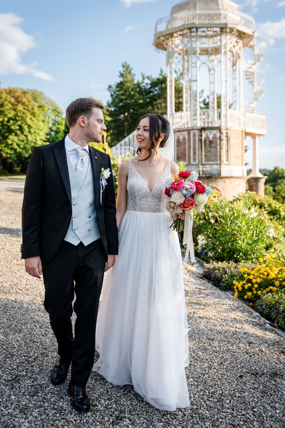 Newlyweds share joyful moments walking together on a gravel path with a decorative gazebo in the background.