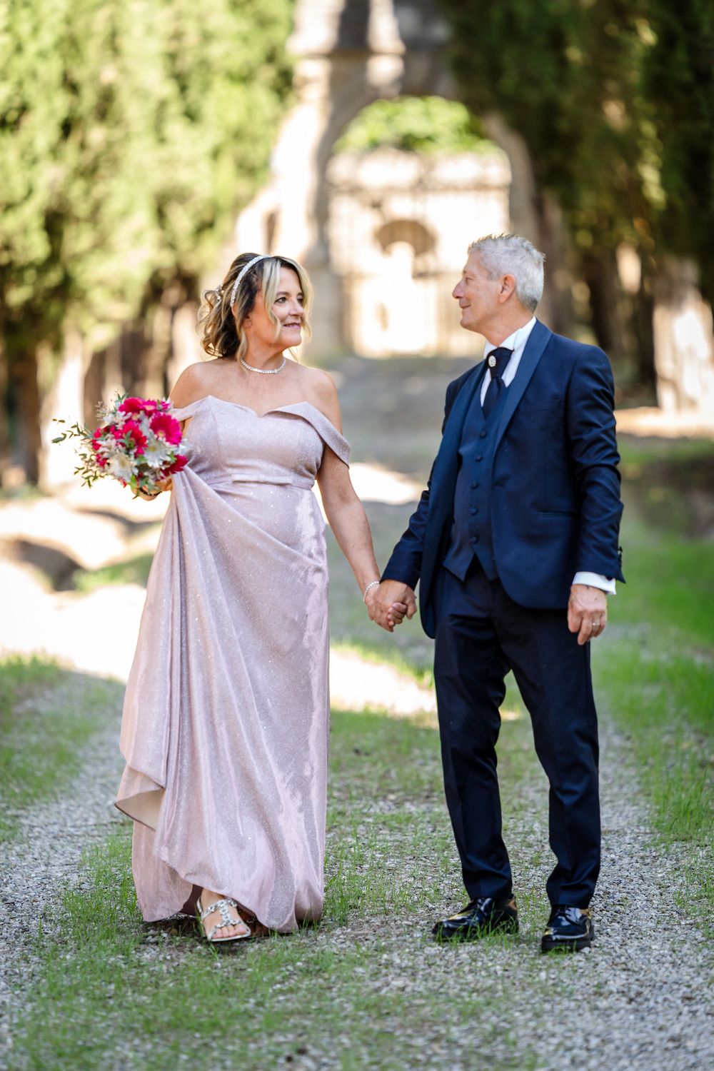 A couple in formal attire dances playfully on a tree-lined path, with the woman in a blush gown and man in navy suit.
