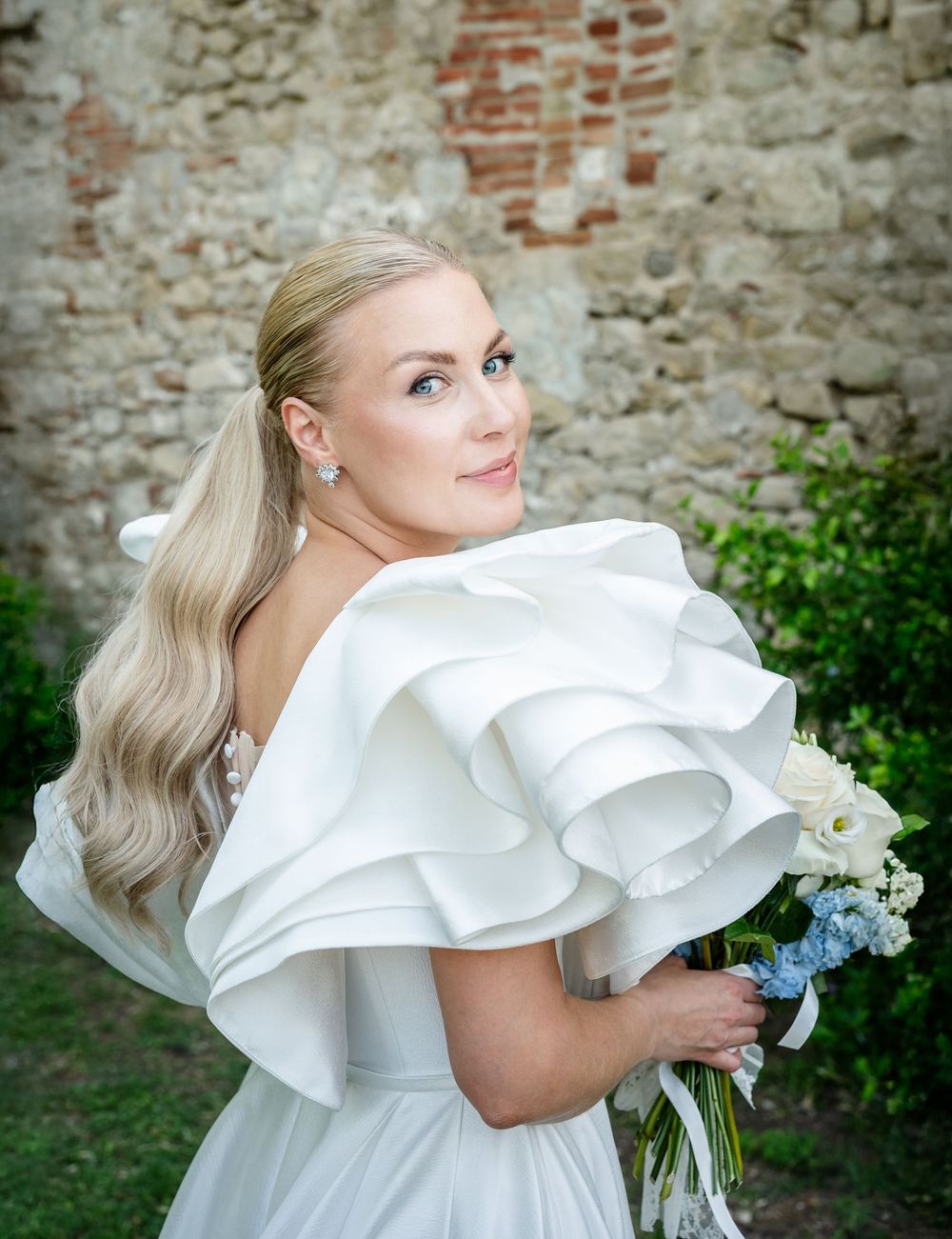 A woman in an elegant white ruffled dress poses in front of a rustic stone wall with flowers.