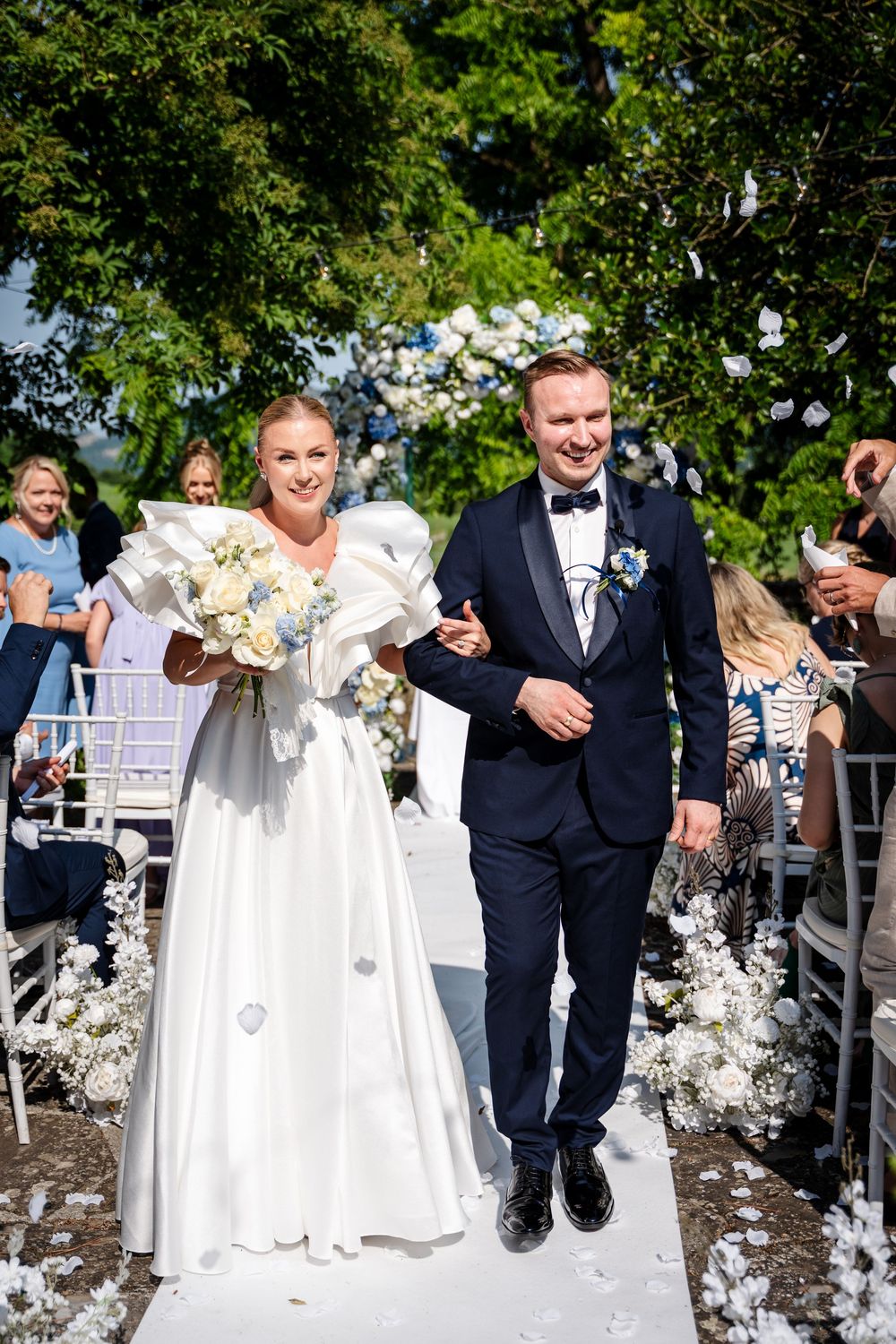 Wedding party joyfully throws white flower petals in an outdoor celebration scene.