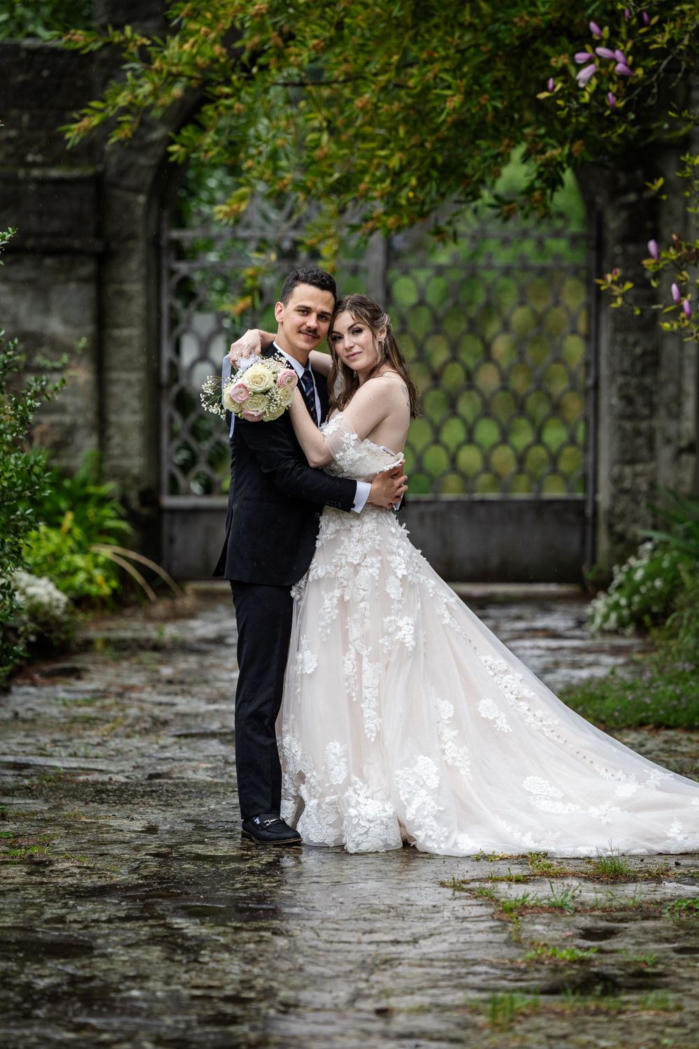 A romantic wedding couple embraces in a garden setting with stone walls and lush greenery on a rainy day.