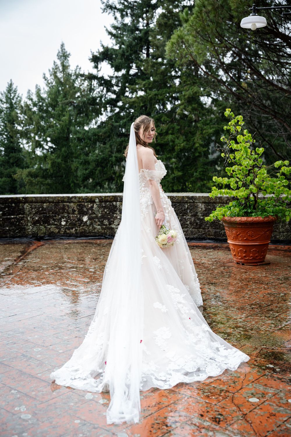 Beautiful bride in a flowing lace wedding dress with train stands on wet brick terrace surrounded by evergreen trees.