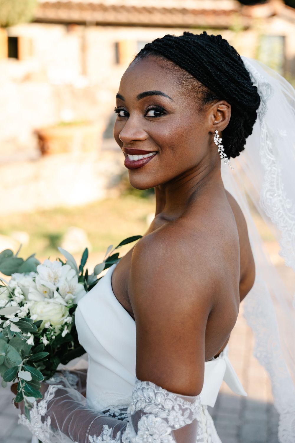 Side profile of bride holding white bouquet with elegant updo hairstyle and lace detailing.