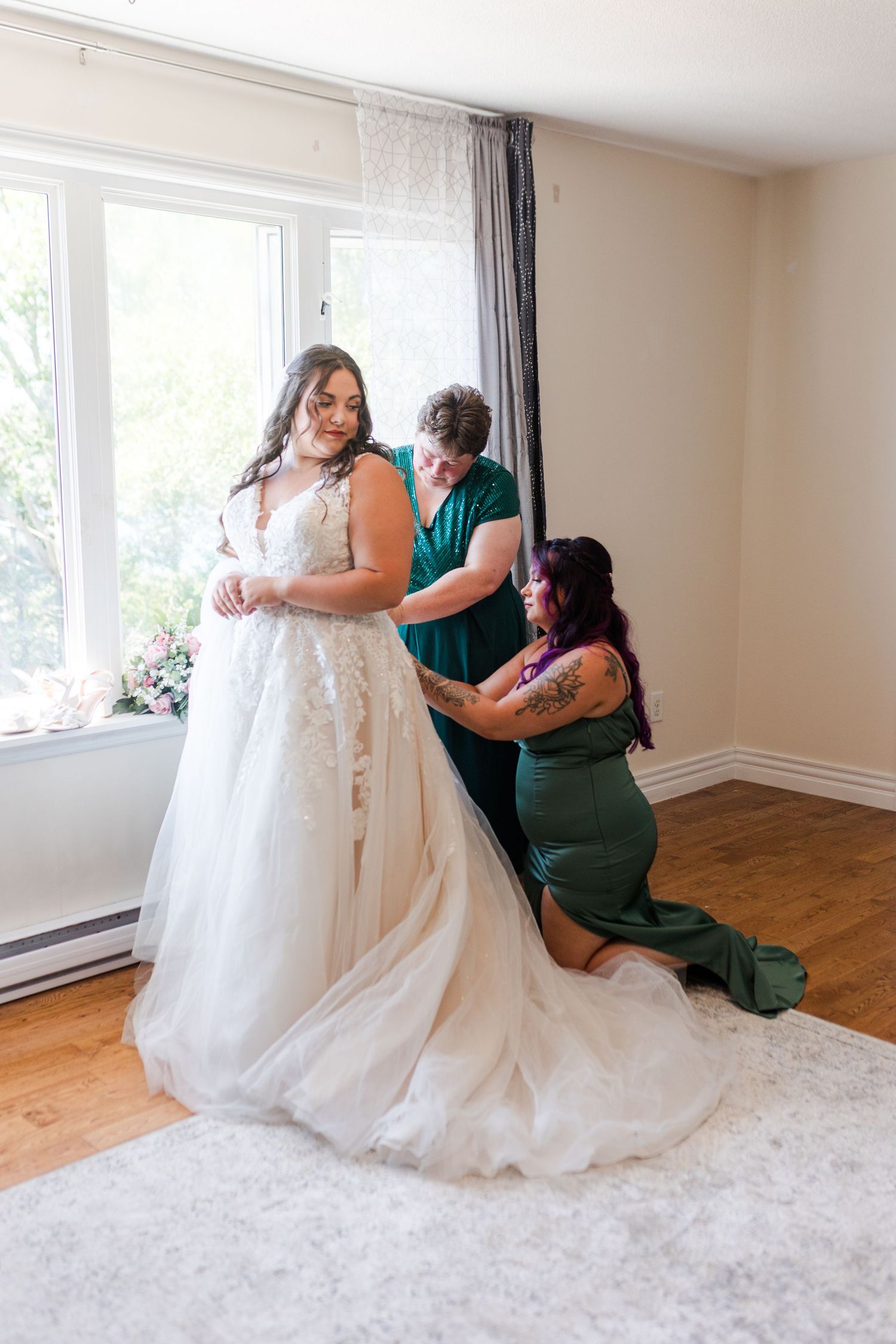 Helpers in emerald green dresses assist with adjusting a flowing white lace wedding gown by a bright window.