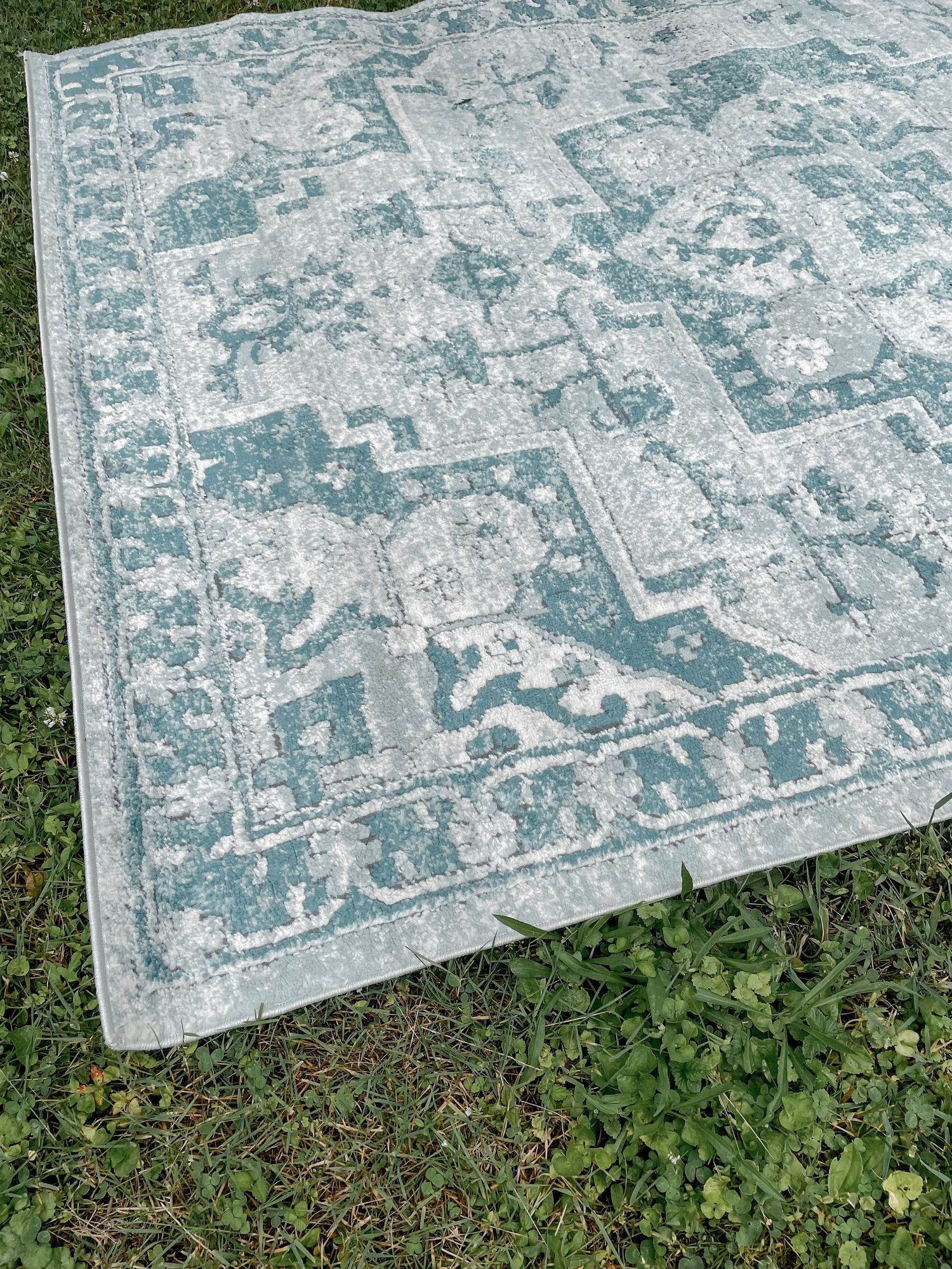 A vintage-style area rug with a distressed teal and white pattern sits partially on grass outdoors.