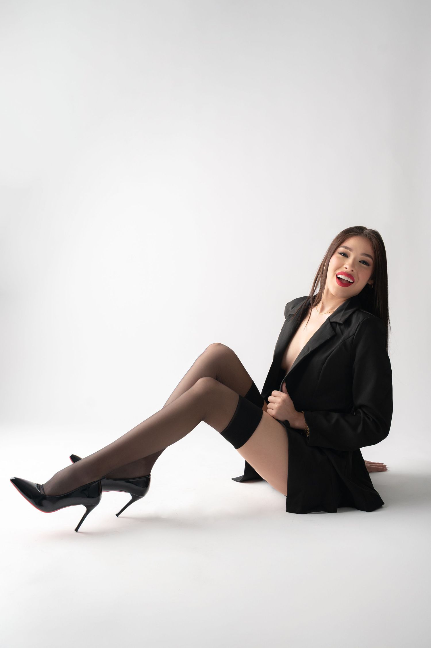 Fashion model in black blazer and stockings poses playfully on white background.