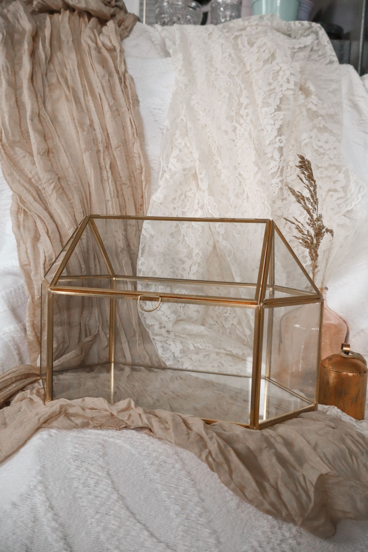 A small glass terrarium house with gold metal frame sits on top of a round rattan side table in a garden setting.