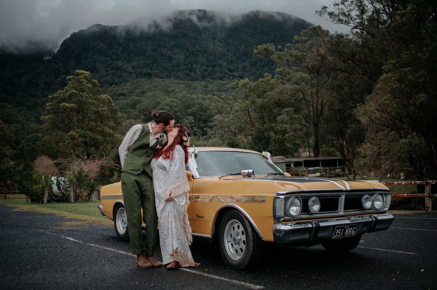 A couple sharing a romantic moment next to a vintage gold Ford XY Falcon GT muscle car with misty mountains in background.