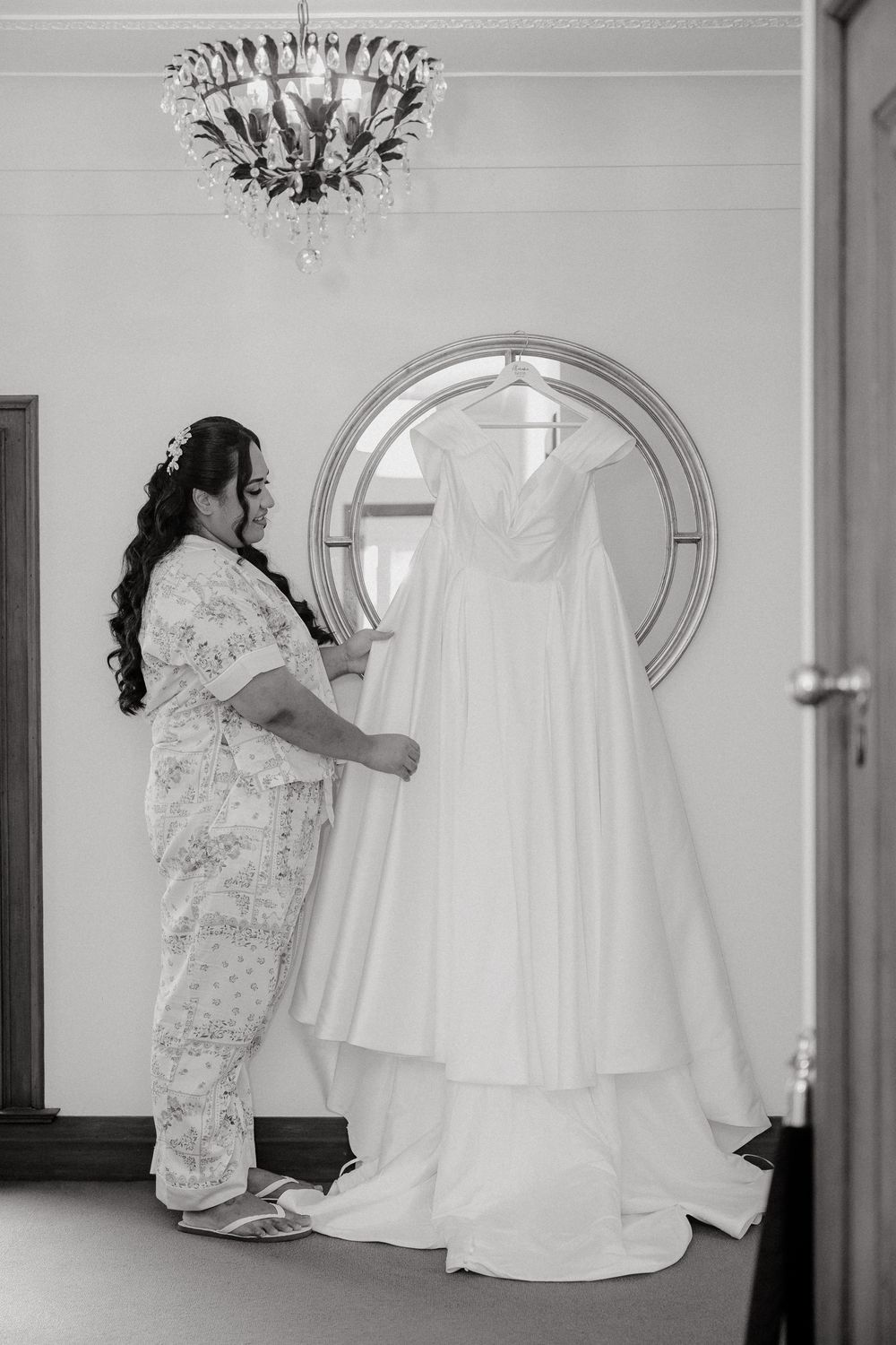Black and white photo showing wedding dress preparations in an elegant room with mirror.