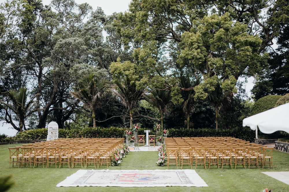 Rows of wooden chairs set up outdoors for a wedding ceremony under tall shady trees.