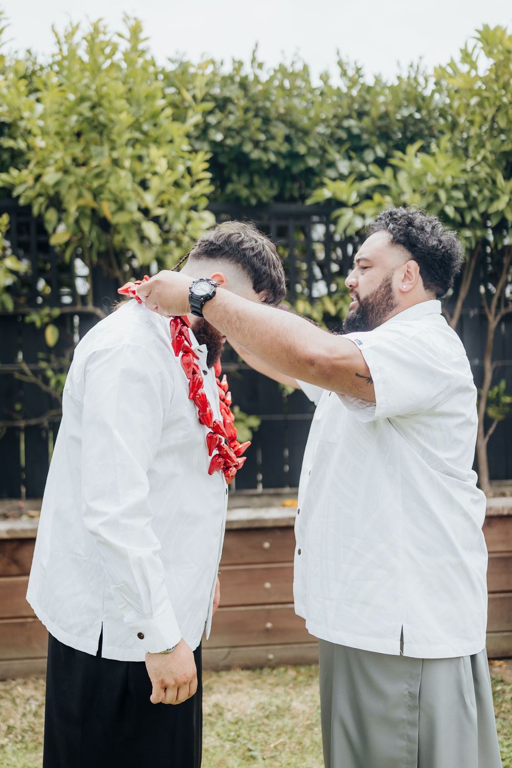 Men in white shirts and traditional Hawaiian leis performing celebratory dance moves outdoors.