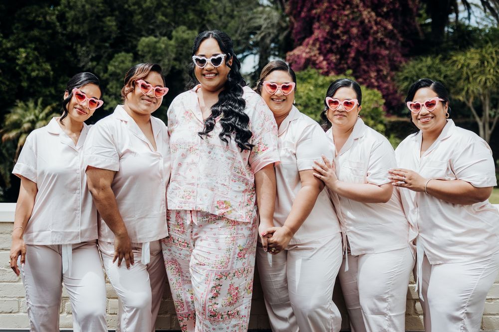 Group photo of wedding party members wearing coordinated white outfits and sunglasses outdoors.