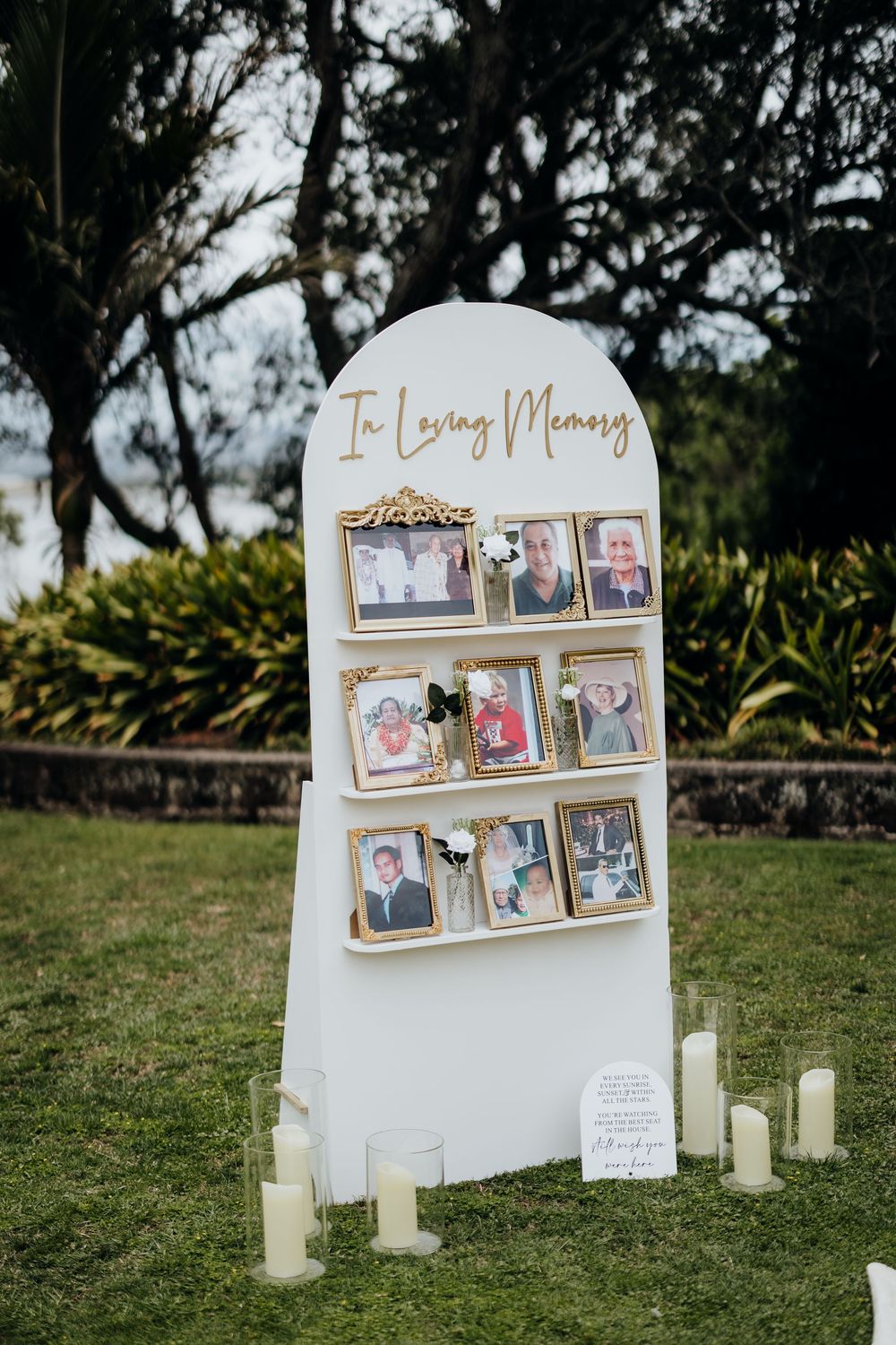 Memorial photo display with vintage pictures on white stands surrounded by candles in an outdoor garden setting.