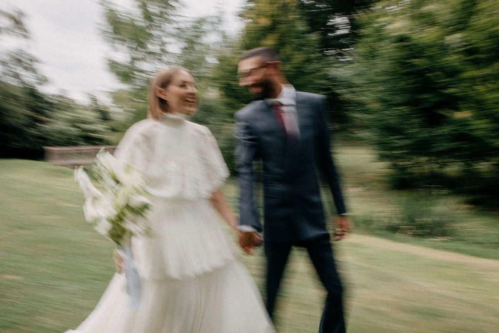 Joyful bride and groom walk hand in hand through lush green gardens in a motion-blur wedding photo.