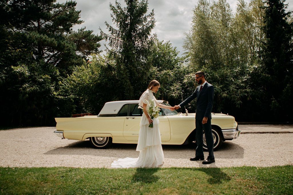 Newlyweds hold hands smiling beside a vintage cream convertible car on a scenic gravel path.