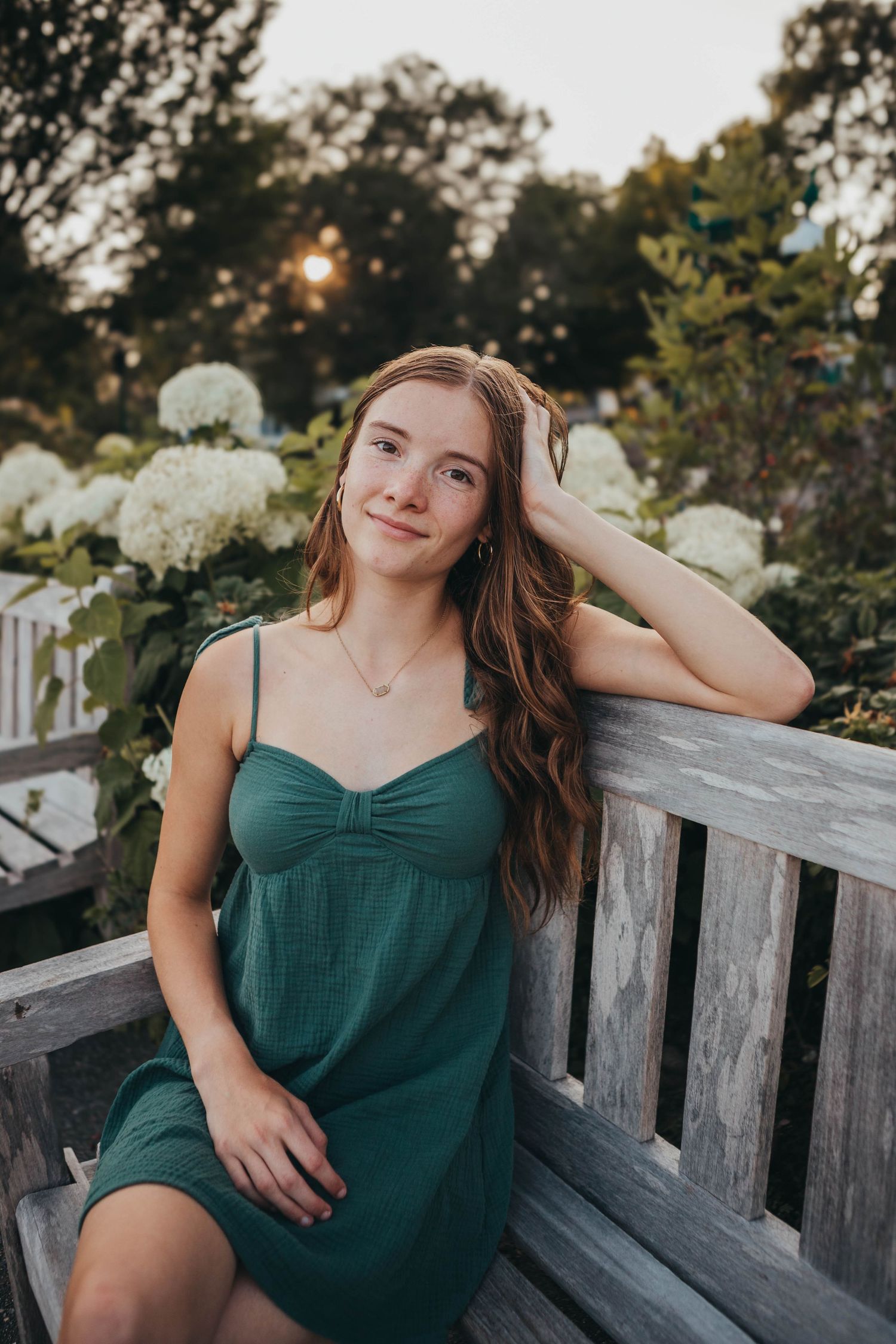 Person in green dress poses casually on wooden bench surrounded by flowers at sunset.