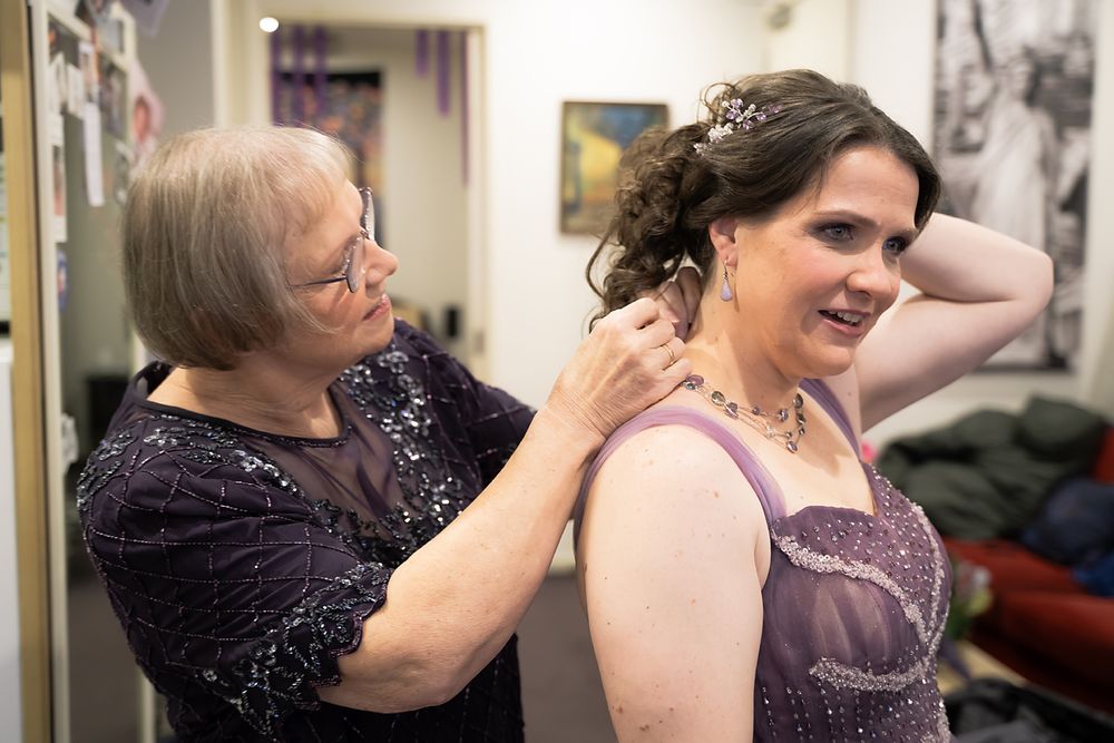 Two people get ready for a formal event, with one helping style the other's hair while wearing elegant evening attire.