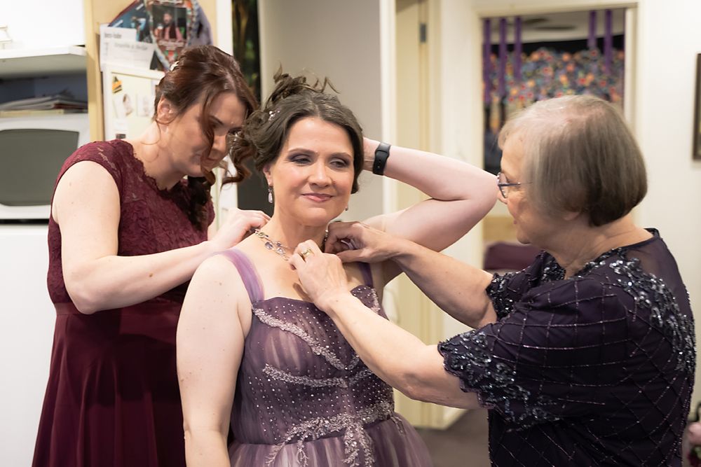 Wedding party members help each other put on corsages and accessories while getting ready in formal attire.