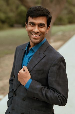A person wearing a charcoal suit and blue shirt poses professionally against a soft outdoor background.