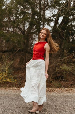 A person in a red top and flowing white skirt twirls playfully outdoors against a natural backdrop.